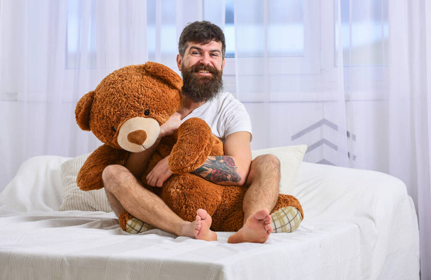 O tipo de cara alegre abraça um urso de peluche gigante. Conceito de aniversário. Homem senta-se na cama e abraça grande brinquedo, cortinas brancas no fundo. Macho com barba e bigode abraçando com brinquedo de pelúcia na manhã de aniversário
 - Foto, Imagem