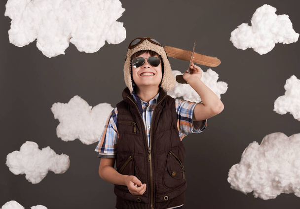 a boy plays with a cardboard airplane and dreams of becoming a pilot, dressed in a retro style jacket and helmet with glasses, clouds of cotton wool, gray background, tinted in brown - Photo, Image