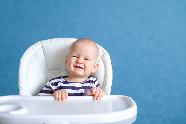 Happy baby. Funny smiling little cutest chubby caucasian white infant child looking at camera on highchair over blue at home - Foto, afbeelding