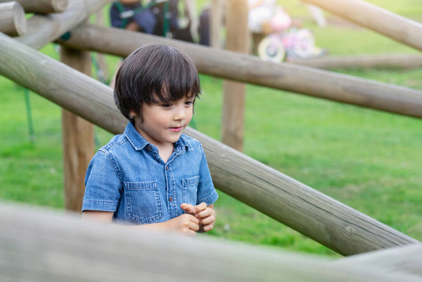 Portret dziecka stojącego z drewnianą sławą w parku, Dziecko korzystające z aktywności w parku przygód wspinaczkowych w letni słoneczny dzień, mały chłopiec bawiący się na placu zabaw - Zdjęcie, obraz