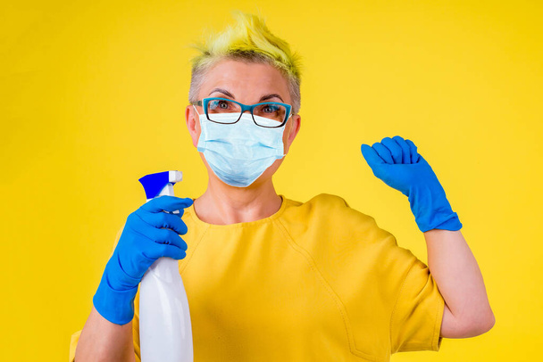 mature woman with colored stylish yellow dyed hair in warm dress and medical mask washing floor at home background studio copy spase - Fotografie, Obrázek