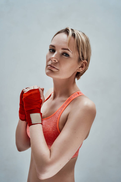 Portrait of a hot and strong woman posing in a bright studio, wearing orange sport bra and hand wraps, while holding her hands prepared for the workout - Photo, Image