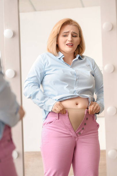 Проблемы с лишним весом женщины в обтягивающей одежде возле зеркала дома
 - Фото, изображение
