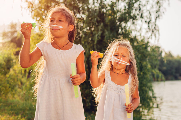 Kislányok húgai buborékokat fújnak a Spring Parkban a folyónál. A gyerekek jól érzik magukat a szabadban. Gyermektevékenységek - Fotó, kép