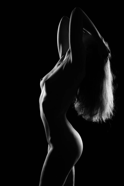 A naked girl straightens her luxuriously long hair as she leans back. Photo of a naked female body in a contoured light on a dark background - Фото, изображение