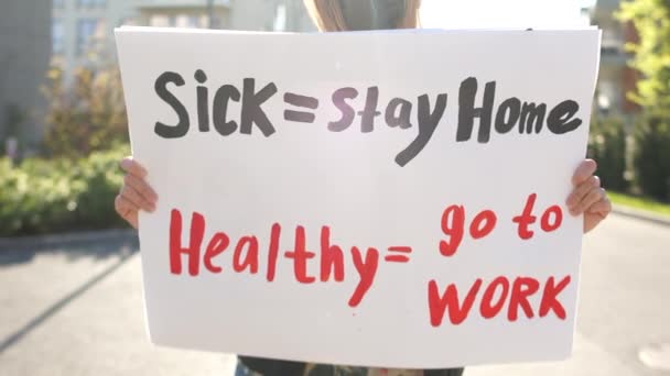 Sick - stay home healthy - go to work. Sunny portrait of a masked girl with a poster against lockdown quarantine restrictions - Materiał filmowy, wideo