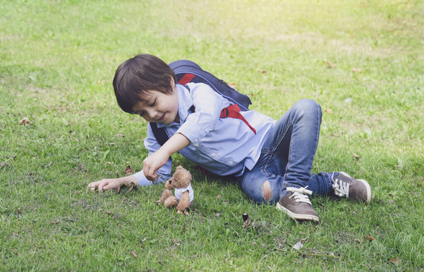 Cute little boy carrying backpack lying on grass playing with teddy bear in te park, Happy kid with smiling face playing with his toy outdoors in spring or summer. - Photo, Image