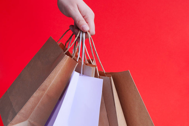 Hand with white paper bags and craft on bright red background. Season of sales or purchases as gift for holidays.Christmas shopping. Courier delivery of products in eco-friendly packaging. copy space - Foto, Imagen