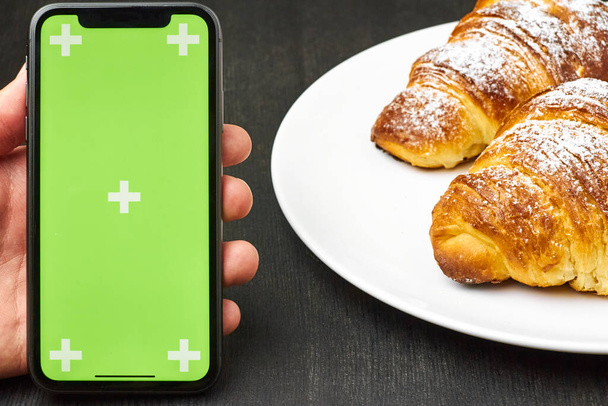 Close up mans hand with a smartphone with a green screen chrome key and croissants on a black background. Croissants with sugar powder on a plate. - Photo, Image