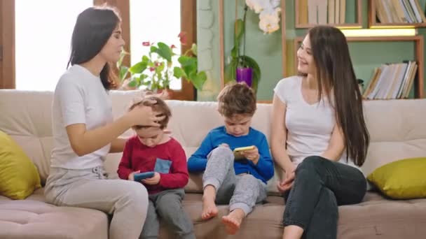 На диване две девушки молодые матери со своими детьми два симпатичных мальчика болтают и улыбаются, пока дети играют в игру на смартфоне остаться дома новый Coronavirus 2019 пандемии
 - Кадры, видео