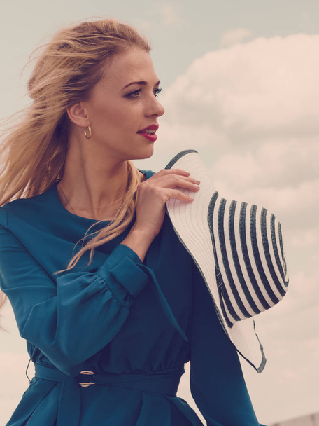 Fashionable woman wearing blue jumpsuit shorts perfect for summer holding elegant stylish sun hat. Fashion model outdoor photo shoot in marine - Φωτογραφία, εικόνα