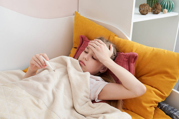 Хвора дівчина-підліток лежить у ліжку і дивиться на термометр
 - Фото, зображення