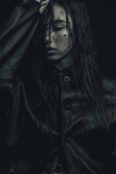 monochrome fashion portrait of a young girl, with wet hair, with drawings on her face, in a leather trench coat, brought her hand to her forehead, half of her face is hidden, sensual lips, large portrait, soft focus - Photo, Image