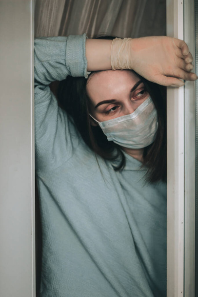 Sairas nuori nainen yllään suojaava kasvonaamio ja käsineet katselee ulos ikkunasta kotinsa koska epidemia korona virus covid-19. Kotikaranteeni ja eristäytyminen
. - Valokuva, kuva