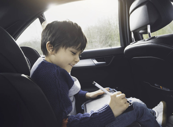 Gelukkig jong jongen met behulp van een tablet computer terwijl zitten in de achterste passagiersstoel van een auto met een veiligheidsgordel, Kind jongen tekening op slimme pad - Foto, afbeelding