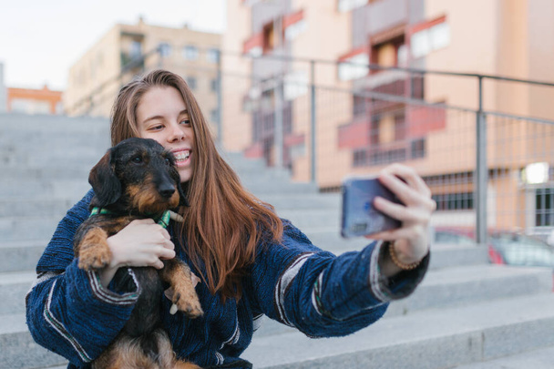 Alternative pretty young adult woman with blue jacket making a selfie photo with her dog outdoors in the city - Fotó, kép