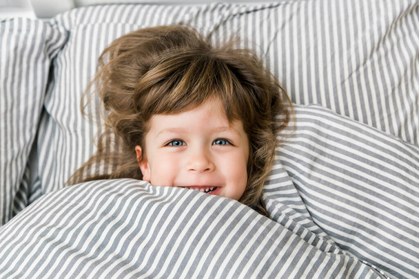 ベッドの中で大きな青い目の子供の少女の肖像画。小さな女の子はにっこりとベッドで楽しんでいます。面白い隔離室の隔離。COVID 19 - 写真・画像