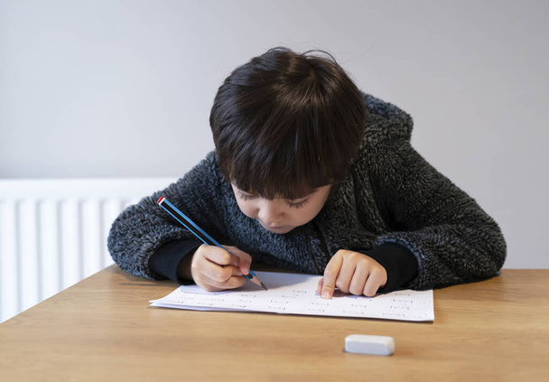 Portrét školního chlapce sedícího na stole, jak dělá domácí úkoly, Happy Child drží tužku psaní, Chlapec psaní anglických slov na bílém papíru, Základní škola a domácí výuka koncepce - Fotografie, Obrázek