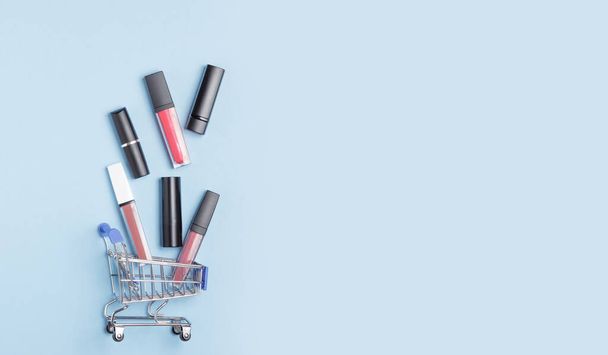 Γυναικεία κραγιόν και lip gloss σε ένα καροτσάκι αγορών σε μπλε φόντο. Η έννοια της αγοράς καλλυντικών, online αγορές. - Φωτογραφία, εικόνα