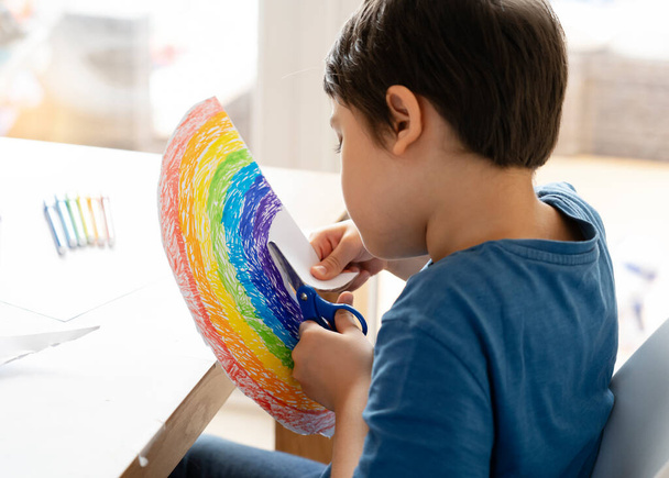  El niño está aislado usando tijeras que cortan papel en forma de arco iris sobre fondo blanco, actividades infantiles en casa mientras está fuera de la escuela.Quédate en casa Campaña de redes sociales para el concepto de prevención del coronavirus
 - Foto, imagen