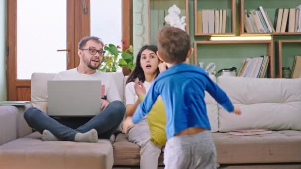 Atrakcyjny szczęśliwy młodzi rodzice spędzają czas na kanapie ich do dzieci wchodzących do salonu szczęśliwy zaczynają walczyć z poduszki uśmiechają się duże - Materiał filmowy, wideo