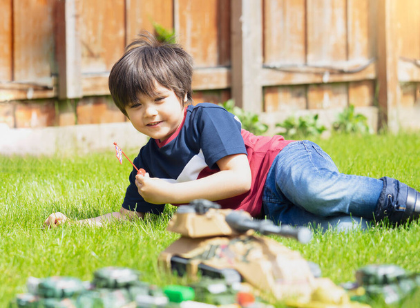 Actieve jongen liggend op het gras spelen met soldaten en tank speelgoed in de tuin, Kid spelen oorlogen en vrede op zijn eigen in hete zonnige dag zomer, Kinderen verbeelding en ontwikkeling  - Foto, afbeelding