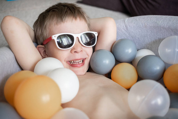 toddler boy in sunglasses swimming in ball pool at home. Self isolation quarantine entertainment at home. Holiday tourism in home - Фото, зображення