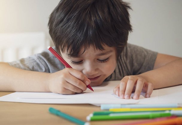 Selektiver Schwerpunkt des Schulkindes, das auf dem Tisch sitzt und Hausaufgaben macht, Happy Child mit rotem Stift schreibt oder auf weißem Papier zeichnet, Grund- und Heimschule, Bildungskonzept - Foto, Bild