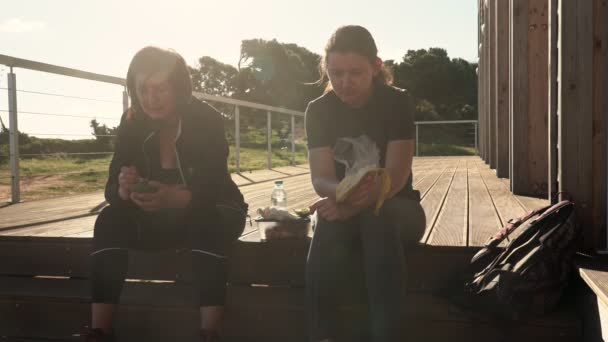 Een succesvolle, gelukkige vrouw van middelbare leeftijd en een jong meisje, moe van een sportieve wandeling, gingen zitten om wat te eten op een houten trap. - Video