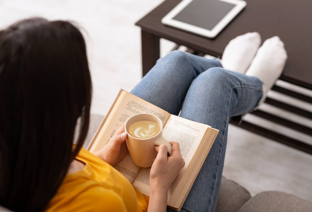 Unrecognizable girl holding cup of fresh aromatic coffee and reading interesting book at home - Zdjęcie, obraz