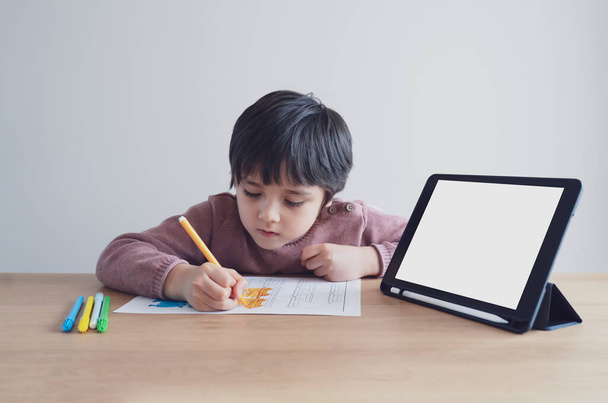Дошкольный ребенок с помощью планшета для его домашней работы, ребенок носить наушники делать домашнее задание с помощью цифровой планшет поиск информации в Интернете, домашнее образование концепции, социальное дистанцирование
 - Фото, изображение
