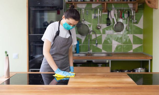 Frau mit Maske und Handschuhen putzt elektrische Keramikherdplatte in der Küche - Foto, Bild