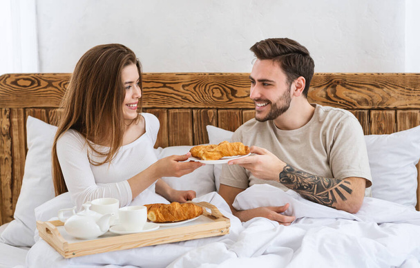 Romanttinen aamiainen kahdelle käsitteelle. Tyttö antaa croissantin miehelle, sängyssä
 - Valokuva, kuva