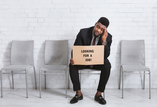 Uomo afro-americano in giacca e cravatta con cartello in cerca di lavoro in attesa di un colloquio in ufficio. Copia spazio
 - Foto, immagini