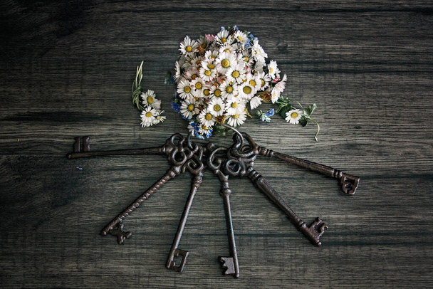 Vintage keys and bouquet of wild flowers in little vase on wooden backdrop - Photo, Image