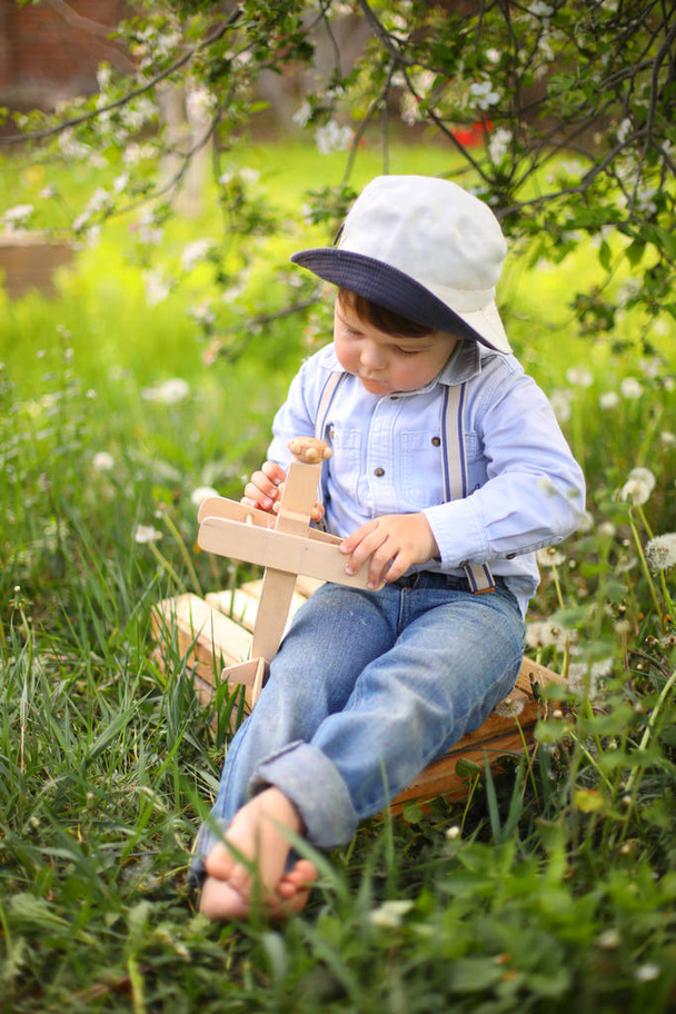 Little cute blond boy playing with a wooden plane in the summer park on the grass on a sunny day - Photo, image