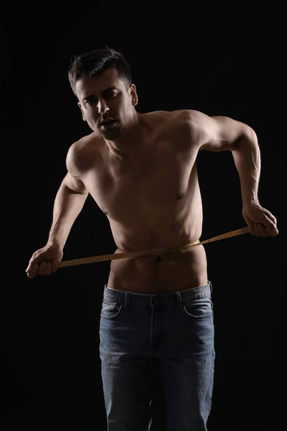 Sick man measuring his waist on dark background. Concept of anorexia - 写真・画像