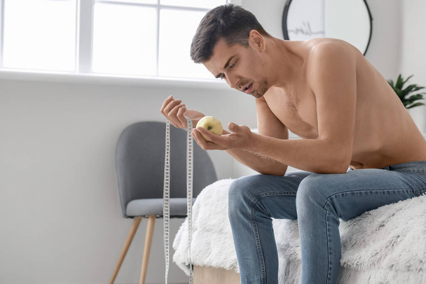 Young man suffering from anorexia with measuring tape and apple in bedroom - Photo, Image