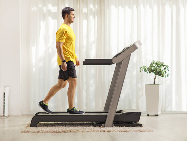 Full length profile shot of a young man walking on a treadmill at home - Zdjęcie, obraz