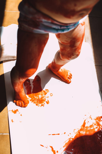 Girl paints a white canvas with paint, dirtying her hands and feet with red paint. - Photo, Image