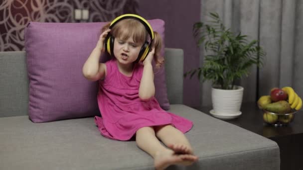 Kleines Mädchen mit Kopfhörern, das Musik hört. Zu Hause auf dem Sofa tanzen - Filmmaterial, Video