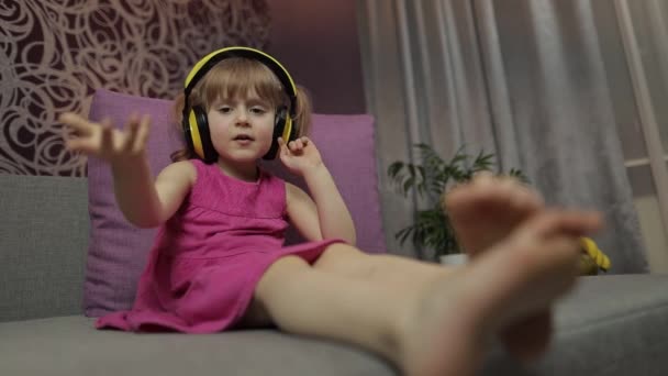 ヘッドフォンで音楽を聞いて楽しむ小さな子供の女の子。自宅でソファで踊る - 映像、動画