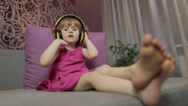 Little child girl in headphones enjoying listen music. Dancing on sofa at home - Footage, Video
