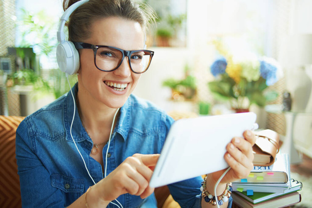 Портрет щасливої стильної домогосподарки середнього віку в джинсовій сорочці з білими навушниками та планшетними комп'ютерами, які вивчають онлайн в сучасному будинку в сонячний день
. - Фото, зображення