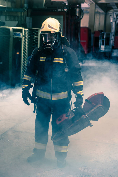 Retrato de un bombero con un equipo de protección completo posando con una motosierra en el hombro. Fondo oscuro con humo y luz azul. - Foto, imagen