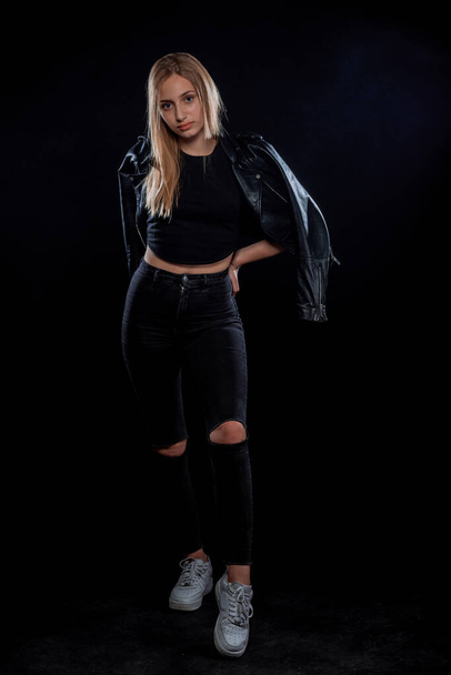 Stylish young woman wearing black leather jacket and white jeans isolated on black background - Fotó, kép