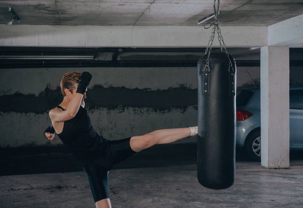 Белая женщина в спортивной одежде и с черными бинтами пинает боксерскую сумку в гараже - Фото, изображение