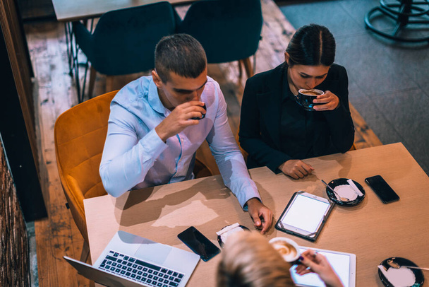Bovenaanzicht van een mooi jong echtpaar dat met hun makelaar praat terwijl ze tablet gebruiken en koffie drinken aan tafel in café - Foto, afbeelding