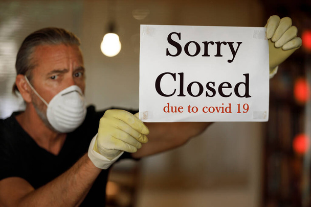coronavirus closed for business concept, male business owner with medical mask puts sorry closed sign on window during covid 19 virus pandemic, cafe shop bankrupt, covid-19 restrictions and lockdowns - Photo, Image