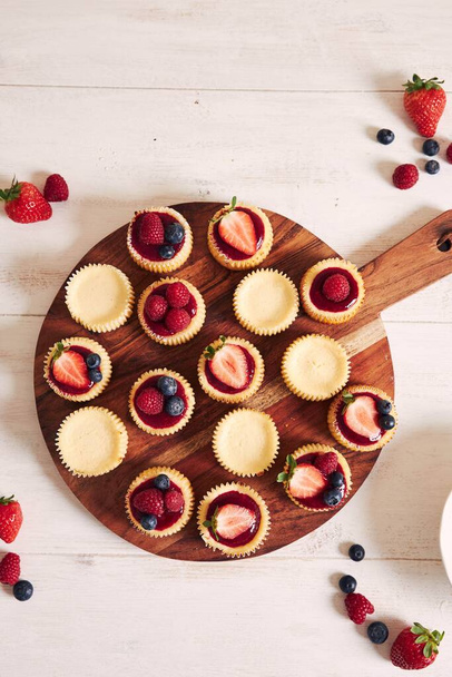A high angle shot of cheese cupcakes with fruit jelly and fruits on a wooden plate with a white background - Фото, зображення